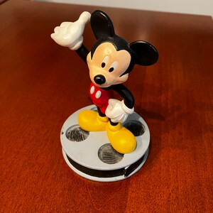May include: A black and red Mickey Mouse figurine standing on a film reel base. Mickey is wearing yellow shoes and has a white glove on his right hand. He is giving a thumbs up.