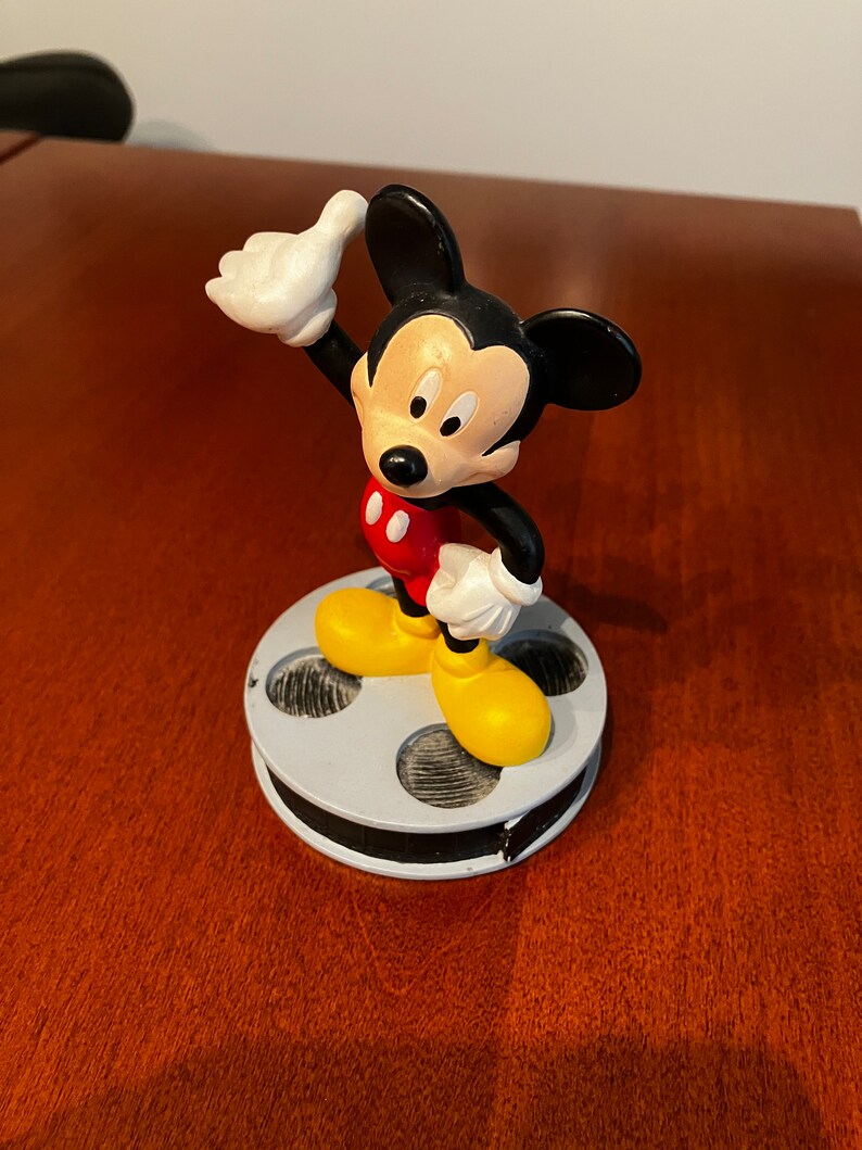 May include: A black and red Mickey Mouse figurine standing on a film reel base. Mickey is wearing yellow shoes and has a white glove on his right hand. He is giving a thumbs up.