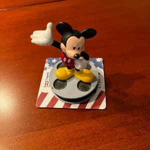 May include: A small plastic figurine of Mickey Mouse waving with a red and yellow outfit standing on a film reel base with an American flag design.