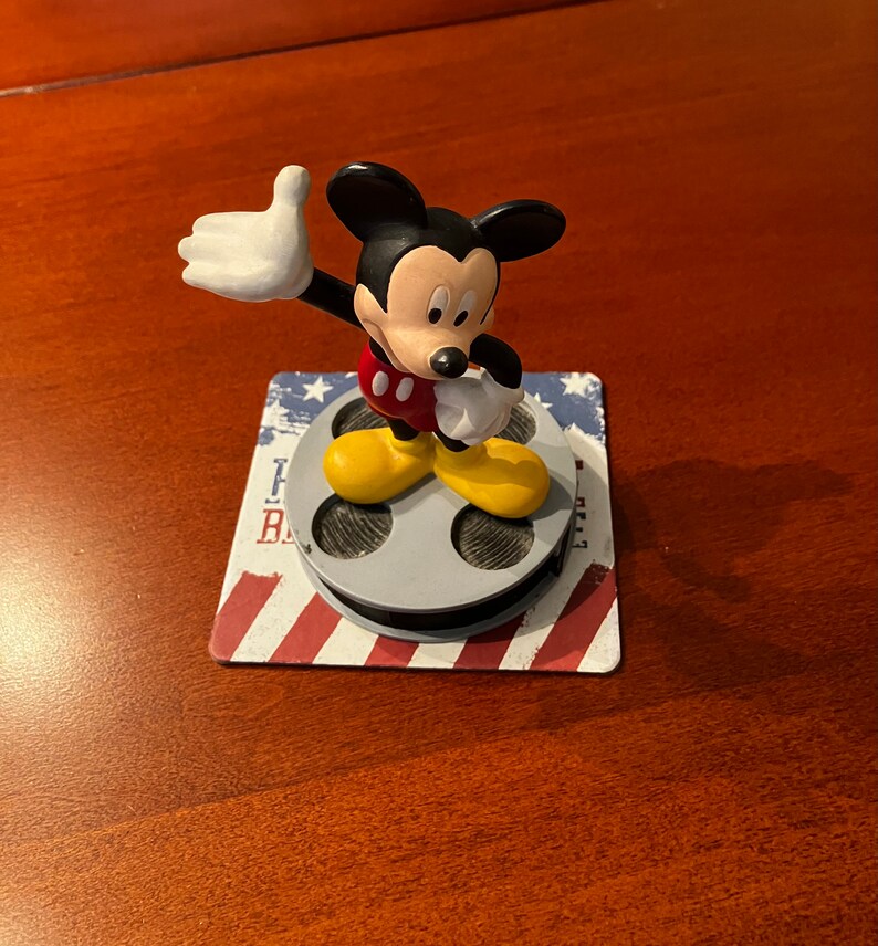 May include: A small plastic figurine of Mickey Mouse waving with a red and yellow outfit standing on a film reel base with an American flag design.