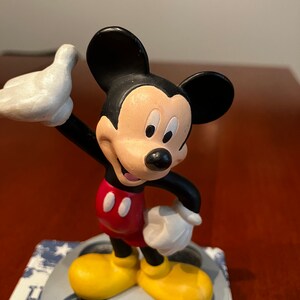 May include: A black and white Mickey Mouse figurine standing on a film reel base. Mickey is wearing red shorts with two yellow buttons and yellow shoes. He is waving with his right hand.