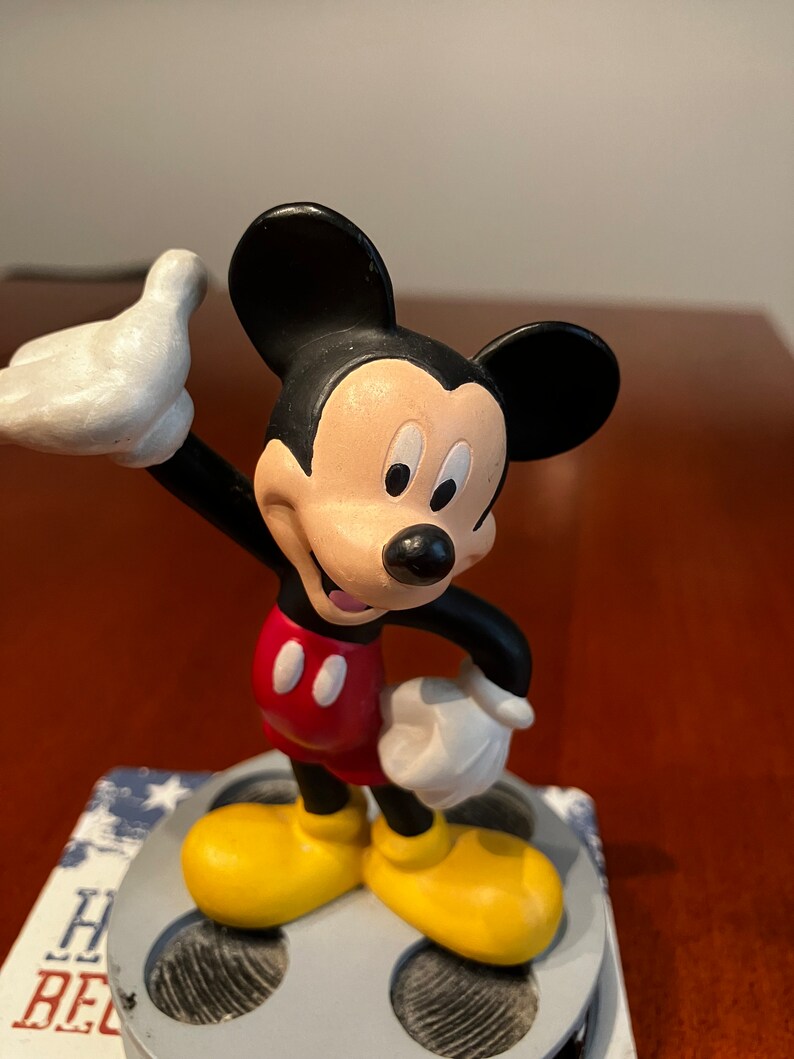 May include: A black and white Mickey Mouse figurine standing on a film reel base. Mickey is wearing red shorts with two yellow buttons and yellow shoes. He is waving with his right hand.