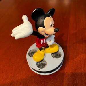 May include: A black and white Mickey Mouse figurine standing on a film reel base. Mickey is wearing red shorts and yellow shoes. He is waving with his right hand.