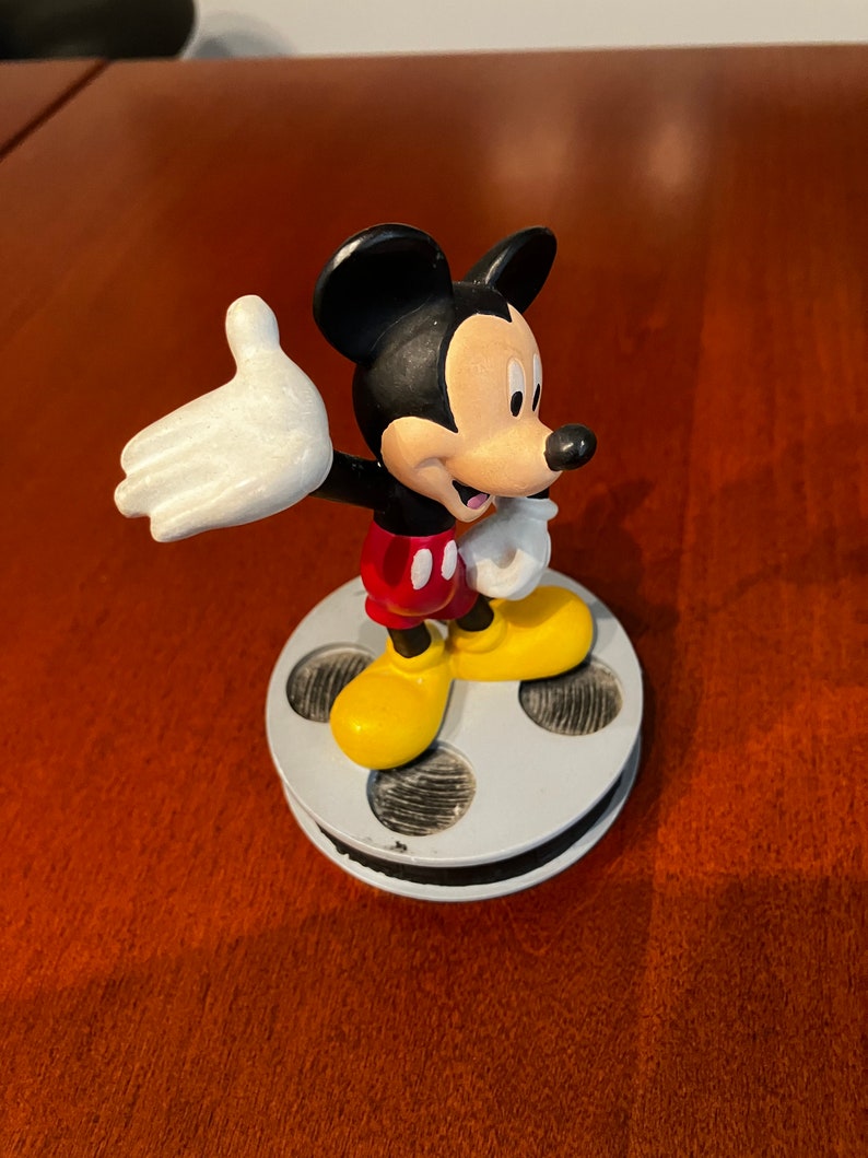May include: A black and white Mickey Mouse figurine standing on a film reel base. Mickey is wearing red shorts and yellow shoes. He is waving with his right hand.