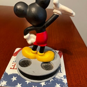 May include: A black and yellow Mickey Mouse figurine standing on a film reel base. The figurine is waving with its right arm and has a red shorts.