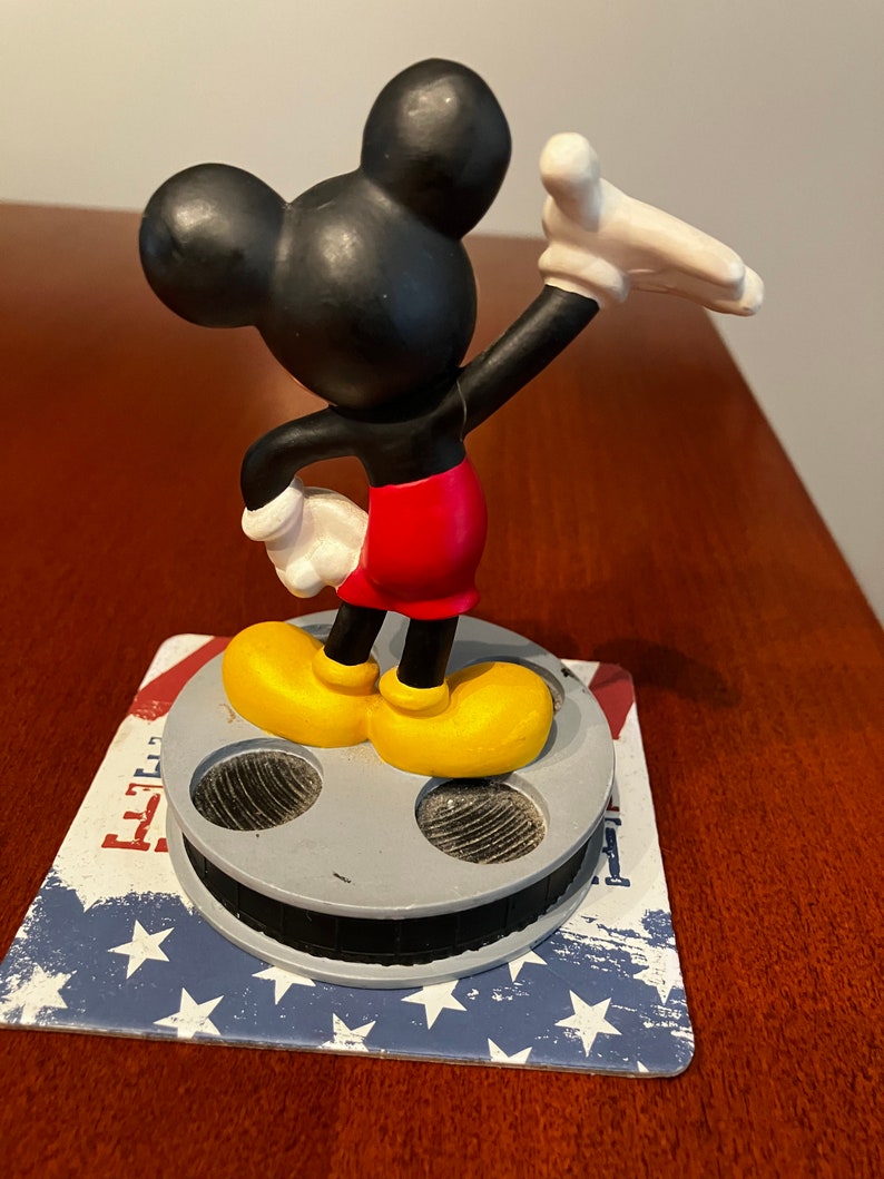 May include: A black and yellow Mickey Mouse figurine standing on a film reel base. The figurine is waving with its right arm and has a red shorts.
