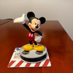 May include: A red and black Mickey Mouse figurine standing on a film reel base with a red, white, and blue background. The base says "Hollywood" and "Believe".