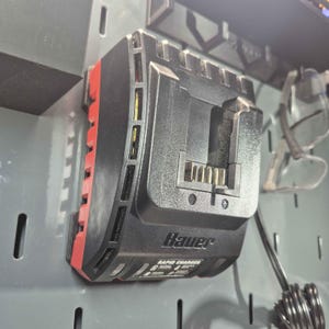 Pode incluir: Carregador rápido Bauer preto e vermelho montado numa placa perfurada cinzenta. O carregador tem uma ranhura para uma bateria e várias luzes indicadoras. O texto "Bauer" e "Rapid Charger" é visível no dispositivo.