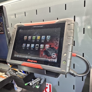May include: A Snap-on Verus Edge diagnostic tablet with a black and red ruggedized frame, featuring a touchscreen display with various software icons, a power button, and a connected black cable, mounted on a gray slotted wall panel.