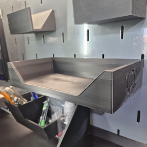 May include: A matte black 3D-printed storage tray with an open front, mounted on a gray pegboard. The side of the tray features a circular logo with the text "DashBoard Gears" embossed on the surface. The tray has a rectangular base and walls.