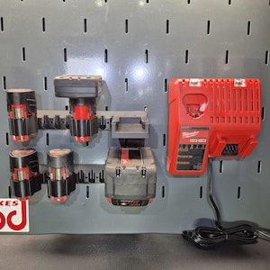 May include: A red and black Milwaukee M12 and M18 battery charger mounted on a gray pegboard, alongside a black plastic battery storage rack holding four red and black power tool batteries of varying sizes and shapes.