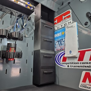 May include: A gray metal cabinet with four drawers is positioned inside a vehicle. The cabinet is next to a pegboard with tools and safety glasses. Stickers with text like "Competition Clutch" and "INKI WINTER" are visible.