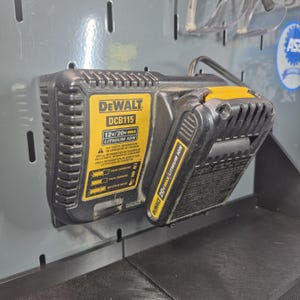 Op de afbeelding: Een zwart-gele DeWalt DCB115 acculader gemonteerd aan een muur, voorzien van een 12V/20V MAX lithium-ion label. Een zwart-gele DeWalt 20V MAX lithium-ion accu is in de lader geplaatst, die is voorzien van oplaadindicator-pictogrammen.
