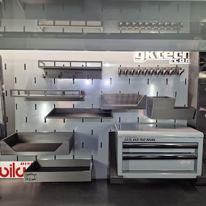 May include: A gray metal pegboard wall organizer system featuring multiple shelves, trays, and a hook rack. The setup includes a small U.S. General tool chest, a shelf labeled Keys/Glasses, and a tray with a DeadPixel 3D Prints logo.