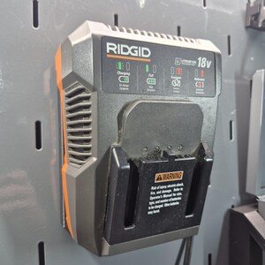 May include: A Ridgid 18v lithium-ion battery charger in a dark gray and orange housing. The front panel features status indicator lights for charging, full, evaluate, and defective states, along with a warning label regarding electric shock risks.
