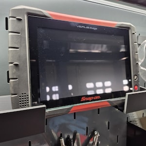 May include: A black and red Snap-on VERUS Edge diagnostic tablet mounted on a black pegboard. The tablet has a large screen and is surrounded by a protective black frame. The Snap-on logo is visible.