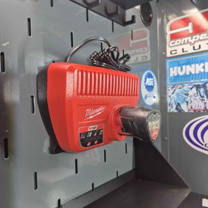 May include: A red and black Milwaukee M12 battery charger mounted on a gray pegboard. The charger has a ribbed design and the Milwaukee logo. A black power cord is connected to the charger.