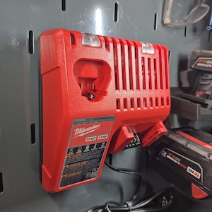 May include: A red Milwaukee M12/M18 battery charger mounted on a gray pegboard. The charger has a rectangular shape with a ribbed top and a charging port. A black battery is partially visible on the right. The charger has a label with safety warnings.