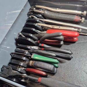 May include: A collection of various metal pliers and locking tools organized in a series of black plastic holders within a drawer. The tools feature handles in red, green, and black, with metallic heads and adjustment knobs visible on the locking pliers.