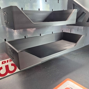 May include: Two black rectangular storage trays mounted on a pegboard wall. Each tray features an open front design with angled side walls and a flat base. The side of each tray displays a small embossed logo with the text "D3D" and "Design".