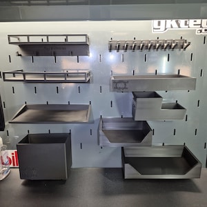 May include: A set of dark gray 3D printed pegboard organizers mounted on a light gray metal panel. The collection includes various shelves, bins, and a hook rack, some featuring the embossed text "RoyalPezol 3D Prints" and "gktech".