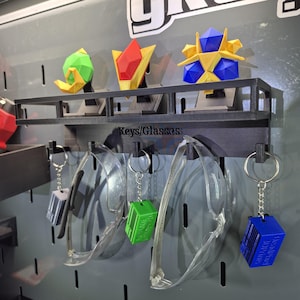 May include: A display featuring geometric keychains and safety glasses. The keychains are in green, blue, and gray, with the text "Keys/Glasses" above the glasses. The keychains are hanging on a pegboard.