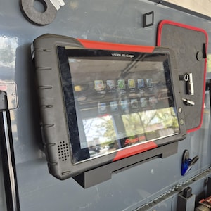 Può includere: Un tablet diagnostico Snap-on VERUS Edge nero e rosso montato su una superficie grigia. Il tablet mostra varie icone di app sullo schermo. Il logo Snap-on è visibile in basso. Un bordo rosso incornicia la parte superiore del tablet.