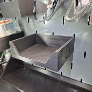 May include: A matte black plastic storage bin with an open front, designed to mount onto a gray pegboard. The side of the bin features a small, embossed circular logo. The bin is rectangular with a low-profile front edge for easy access.