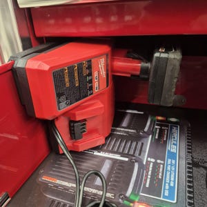 Op de afbeelding: Een rode Milwaukee M12 en M18 acculader horizontaal gemonteerd, met een zwarte accu aan de zijkant bevestigd. Daaronder bevindt zich een zwarte Hercules 20V/12V lithium-ion dubbele lader met een zichtbare USB-poort en gedrukte tekst.
