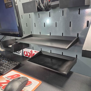 May include: Two black rectangular storage trays mounted onto a gray vertical pegboard wall. The trays feature a flat base with raised edges, designed for organizing small items. The pegboard includes vertical slots for mounting accessories.