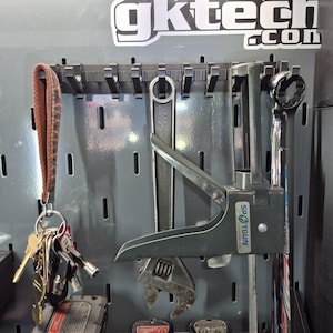 May include: A grey metal pegboard wall organiser with the text gktech.com featuring a brown leather keychain with keys, a large adjustable wrench, a black Spotgun tool, and a chrome wrench hanging from black plastic hooks on the slotted panel.