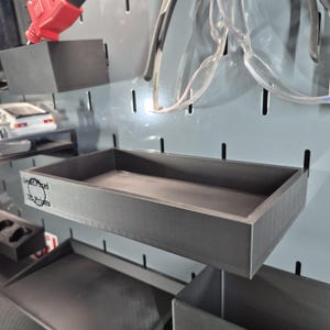 May include: A dark gray rectangular shelf with a low profile, mounted on a pegboard. The shelf has a simple, box-like design. The text "DePixel 3D Prints" is visible on the side. Other shelves and tools are also visible in the background.