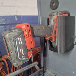 May include: A red and black cordless power tool with an M18 Red Lithium XC5.0 battery attached, mounted horizontally into a black plastic wall-mounted storage bracket. The tool features a textured grip and is secured against a grey metal surface.