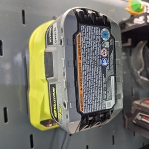 Op de afbeelding: Een Ryobi 18V ONE+ 4Ah lithium-ion accupack met een behuizing van limoengroen en grijs plastic. De accu is voorzien van een opvallend zwart waarschuwingslabel met witte tekst, veiligheidsinstructies, modelnummer PBP004 en UL-certificering.