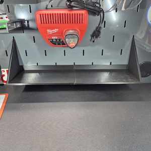 May include: A red Milwaukee M12 battery charger with a black power cord is mounted on a gray pegboard. Below the charger is a black shelf. The pegboard has evenly spaced holes.