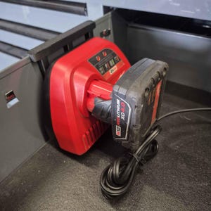 Op de afbeelding: Een rode M12-batterijlader met een zwarte M12 REDLITHIUM XC 4.0-batterij bevestigd. De lader is aangesloten en bevindt zich in een grijze lade.