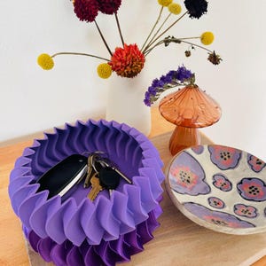 May include: A purple, folded, decorative bowl with a set of keys and a car key inside. The bowl is on a wooden surface next to a small, white and purple patterned dish.