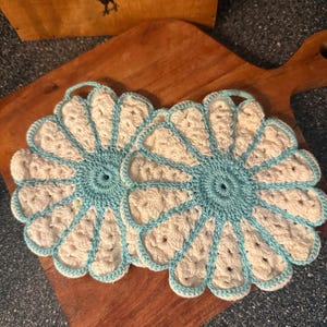 May include: Two round, crocheted potholders shaped like flowers. Each piece features a central circular core in light blue yarn, surrounded by twelve cream-colored petals outlined in the same light blue yarn. A small loop is attached to the top edge.