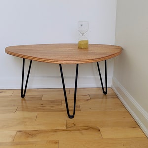 May include: A wooden coffee table with a unique oval shape and black hairpin legs. The table is made of light wood with a natural grain pattern. The table is sitting on a hardwood floor.