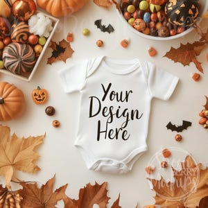 May include: White baby onesie with the text "Your Design Here" on a white background. The onesie is surrounded by fall leaves, candy corn, and small pumpkins.