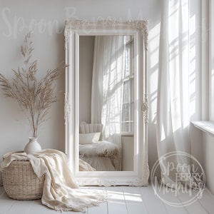 May include: A white ornate mirror with a large, rectangular frame, reflecting a white room with a window and a white chair. The mirror is standing on a white wooden floor with a white throw blanket draped over a wicker basket.