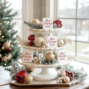 Może przedstawiać: Trójpoziomowy biały stojak na ciasto ozdobiony złotymi obwódkami i pokryty białym lukrem. Stojak jest ozdobiony czerwonymi i złotymi ozdobami, zielenią i czterema białymi kwadratowymi blokami z tekstem "Your Design Here" na każdym.