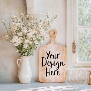 Könnte beinhalten: Ein Holzbrett zum Schneiden mit dem Text "Your Design Here" in schwarzen Buchstaben. Das Brett liegt auf einer weißen Arbeitsplatte neben einer Vase mit weißen Blumen.