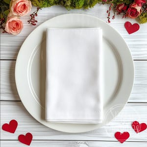 May include: A white folded napkin rests on a white plate, with red heart-shaped cutouts scattered around. Pink roses and greenery are arranged at the top. The scene suggests a romantic or celebratory dining setting.