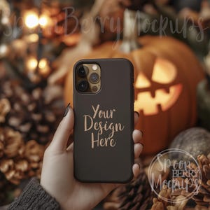 Könnte beinhalten: Eine schwarze Handyhülle mit einem Kameraobjektiv und dem Text "Your Design Here" in goldenen Buchstaben. Die Handyhülle wird von einer Hand vor einem herbstlichen Hintergrund mit Kürbissen und Tannenzapfen gehalten.
