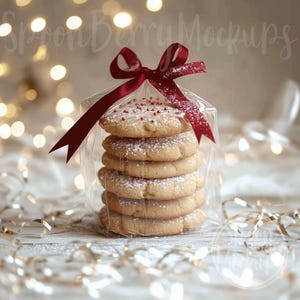 May include: A stack of six sugar cookies tied with a red ribbon and packaged in a clear plastic bag. The cookies are dusted with powdered sugar and have red sprinkles on top. The cookies are sitting on a white surface with gold ribbon and string lights.