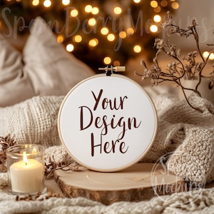 Puede incluir: Un aro de bordado blanco con el texto "Your Design Here" en letras marrones, sentado sobre una rodaja de madera con una manta de punto blanco y una vela encendida en primer plano.
