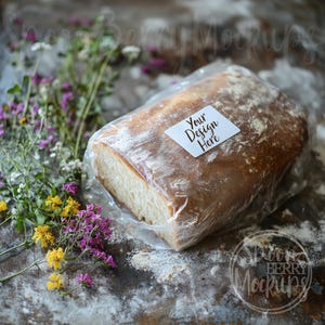 May include: A loaf of bread in a clear plastic bag with a white label that says "Your Design Here" on a rustic wooden surface with scattered flour and wildflowers.