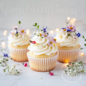 May include: Three white cupcakes with vanilla frosting topped with delicate wildflower arrangements. The cupcakes are on a white surface with string lights in the background.