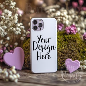May include: White iPhone case mockup with a white background and the text "Your Design Here". The case is sitting on a wooden surface with a pink heart and white flowers.